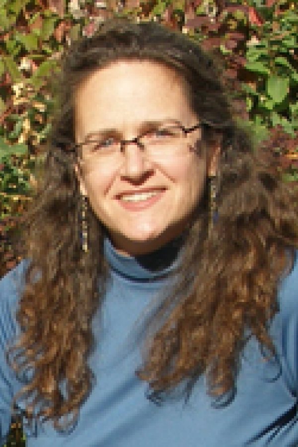 Person with long brown curly hair and glasses wearing a blue shirt against a leafy green background.