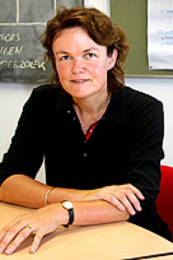 Person in black blazer seated at table in classroom with chalkboard and whiteboard visible in background.