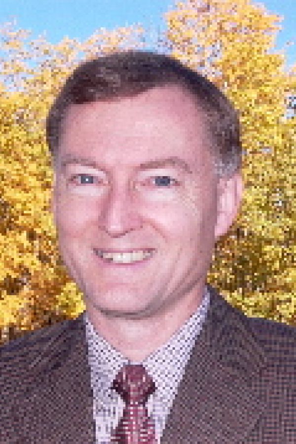 Person smiling at camera wearing brown blazer and patterned tie, with autumn foliage in background.
