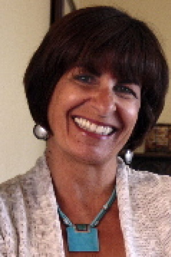 A smiling person with dark hair and bangs wearing a gray cardigan over a teal shirt, indoors.