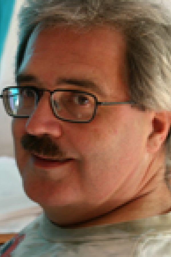 Person with gray hair and glasses wearing a gray shirt against a blue background.