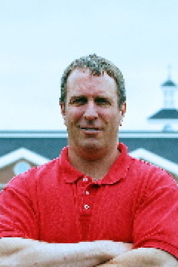 Person in red shirt with arms crossed, standing in front of a cruise ship docked at a port.