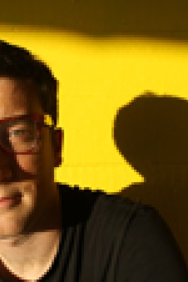 Person wearing glasses and dark clothing against a yellow background with their shadow visible beside them.