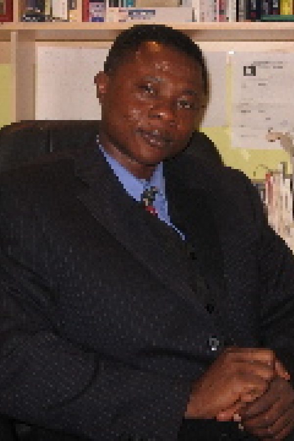 Person in dark suit and blue tie sitting indoors with bookshelves visible in background.