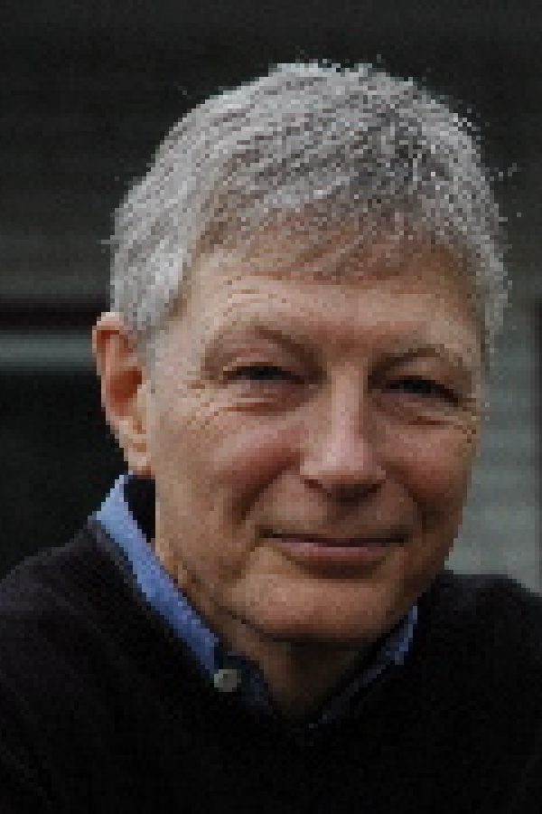 Headshot of an older person with short gray hair wearing a dark jacket over a blue shirt, smiling at the camera against a dark background.