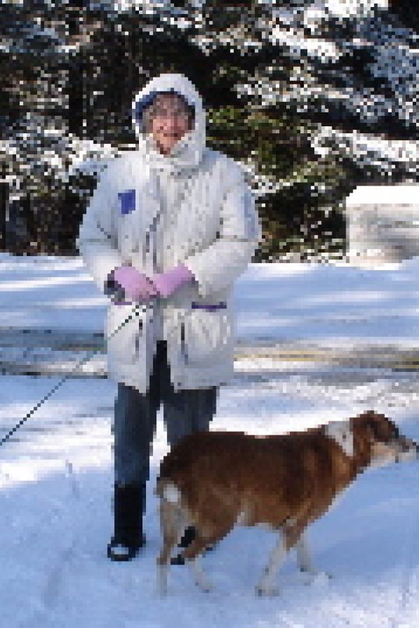 Person in winter clothing standing in snow with a brown dog on a snowy outdoor setting.