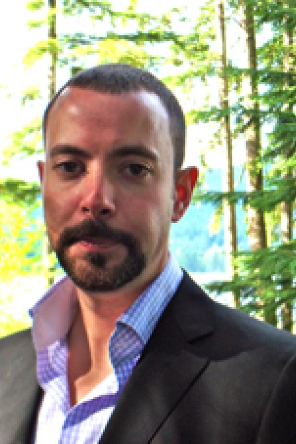Person with dark hair and beard wearing a dark blazer and blue shirt, smiling outdoors with green foliage in background.