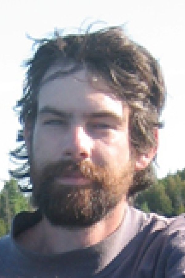 Person with dark curly hair and beard wearing a dark shirt, photographed outdoors with green trees in background.
