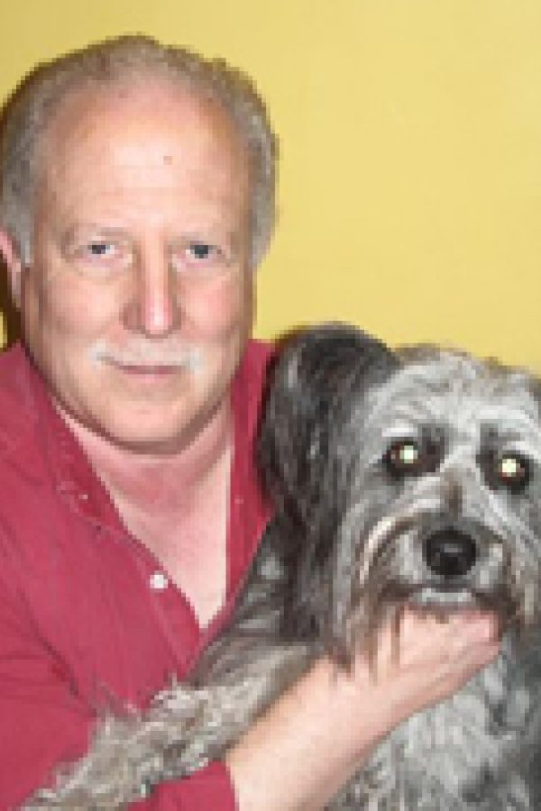 A person in a red shirt holding a gray and white Schnauzer-type dog against a yellow background.