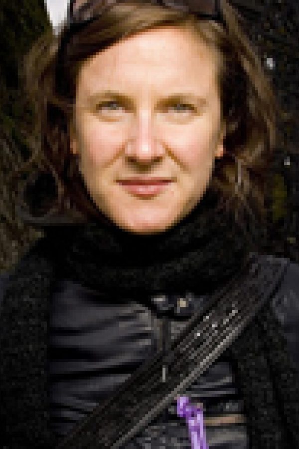 Person with dark shoulder-length hair wearing black clothing against a dark textured background.