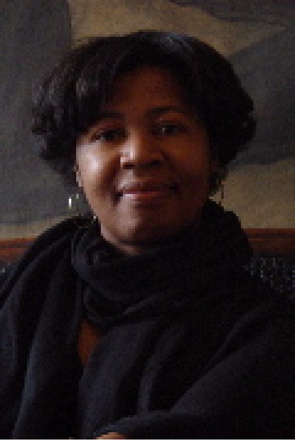 Person with dark curly hair wearing a black jacket and red shirt, smiling at camera against a blurred brown background.