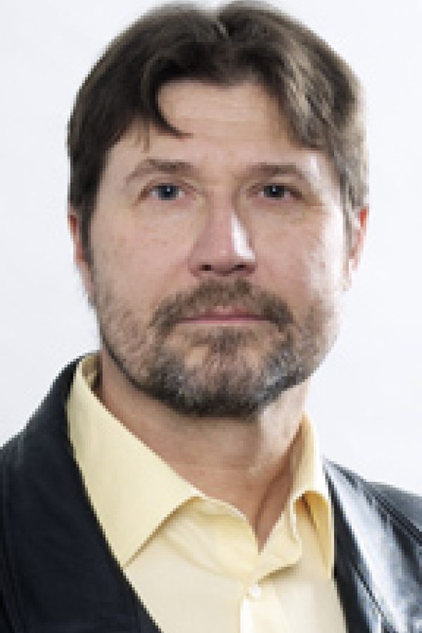 Headshot portrait of an adult person with dark hair and facial hair, wearing a cream-colored shirt and dark jacket against a light background.