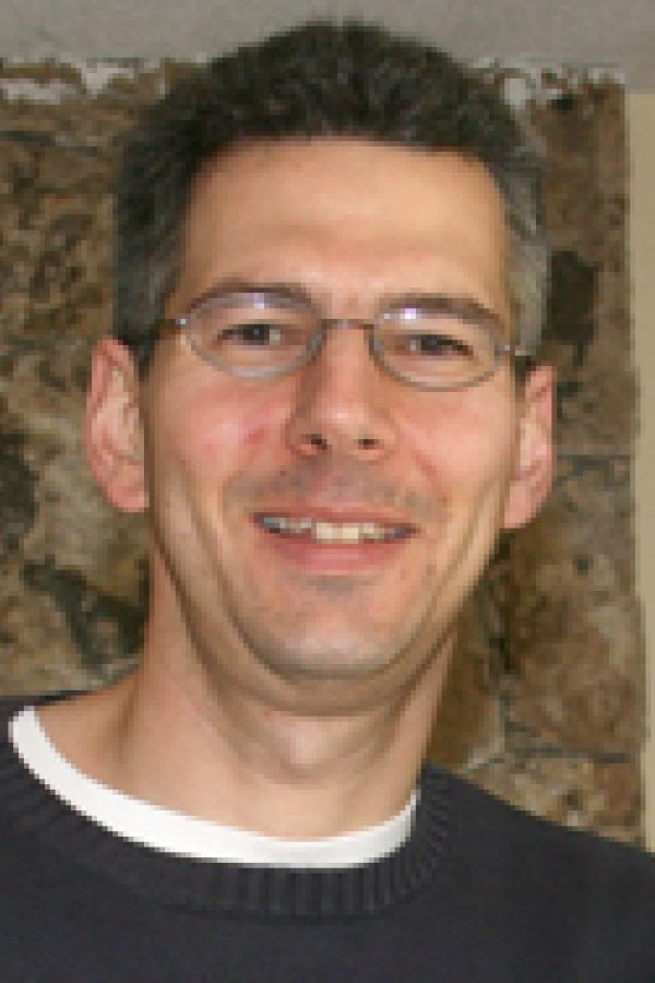 Person wearing glasses and dark shirt smiling at camera against textured beige wall background.