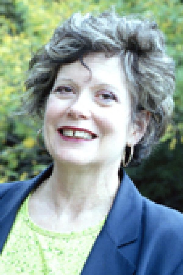 Person with short dark hair smiling at camera outdoors, wearing navy blazer over green top.