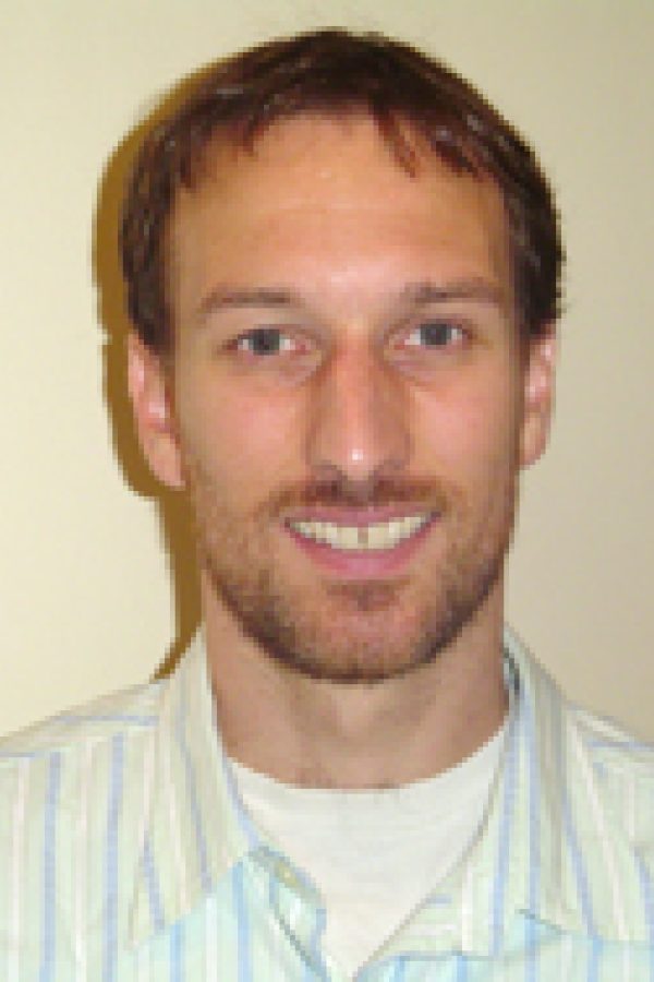 Smiling person with short brown hair wearing a light blue striped shirt against a neutral background.