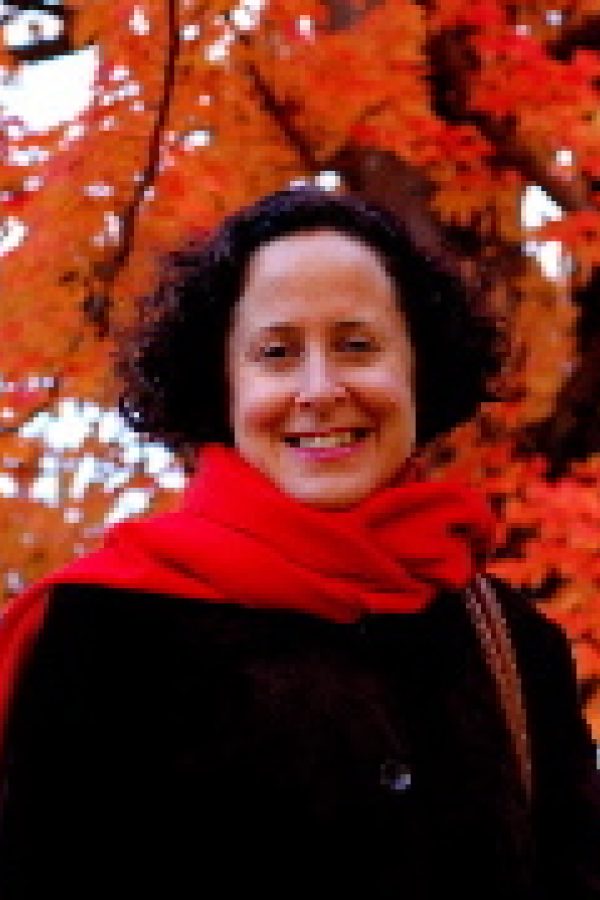 A smiling person with dark hair wearing a red scarf and black jacket stands against a backdrop of vibrant red autumn foliage.