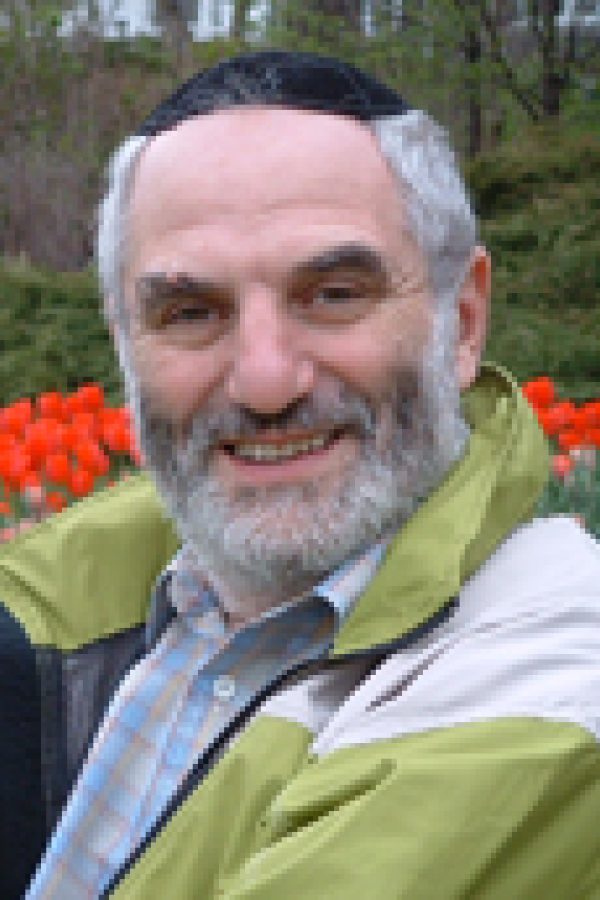 Person with gray beard wearing a yellow-green jacket over blue shirt, smiling outdoors with red flowers in background.