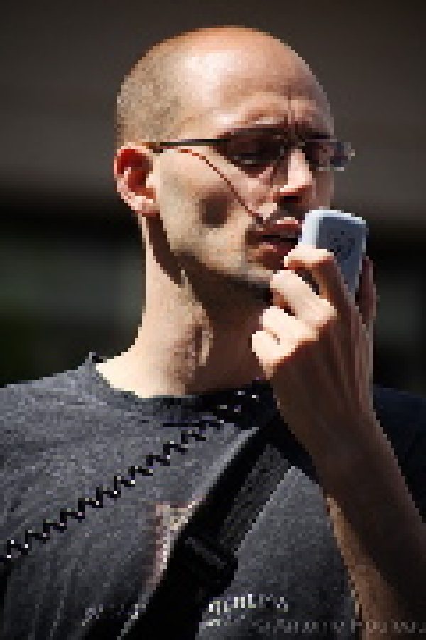 Person wearing glasses and dark clothing speaks into a microphone against a dark background.