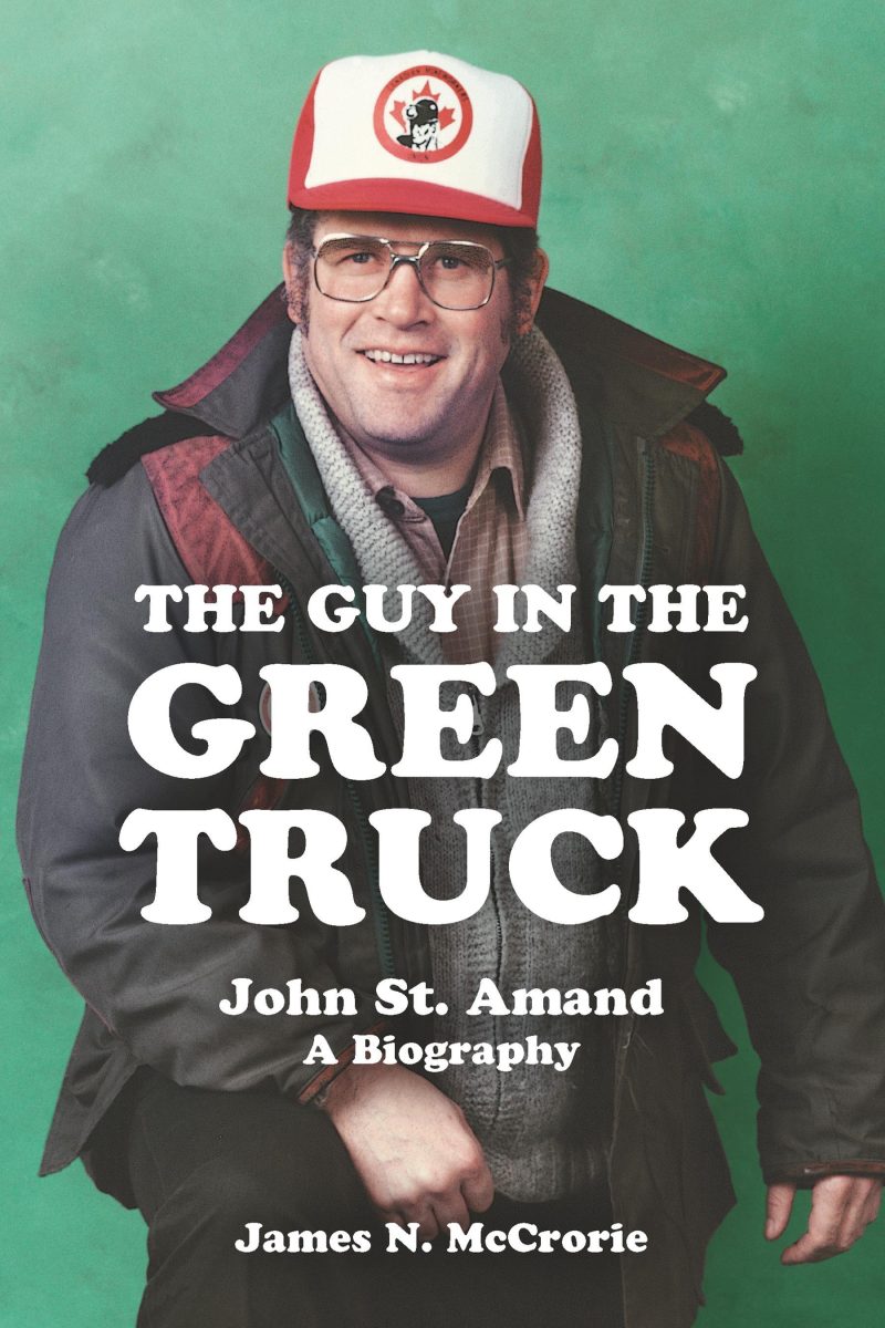 Book cover for "The Guy in the Green Truck" biography by James N. McCrorie about John St. Amand, featuring a person wearing glasses and a red and white cap against a green background.