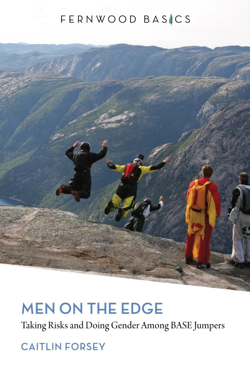 BASE jumpers in colorful gear leap from a cliff edge above a forested valley with mountains in the background.