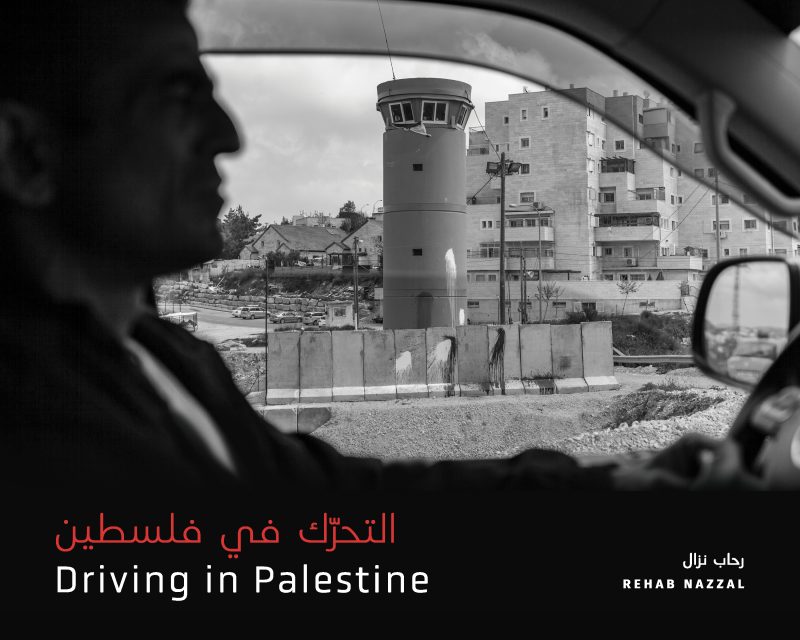 View from inside a car window showing a concrete watchtower and barrier wall with residential buildings in the background, Palestine.