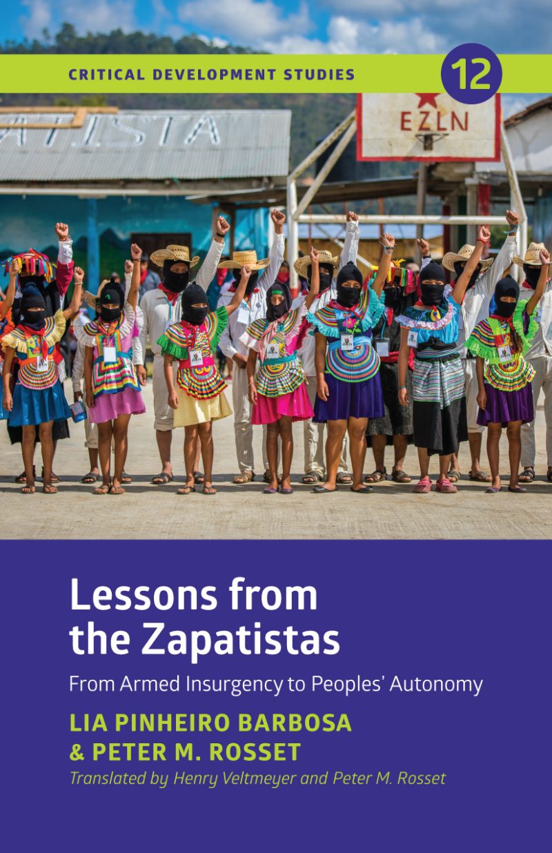 Group of Indigenous people wearing traditional colorful beaded necklaces and clothing, standing with fists raised outside a blue building with EZLN sign.