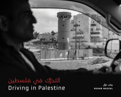 View from inside a car window showing a concrete watchtower and barrier wall with residential buildings in the background, Palestine.