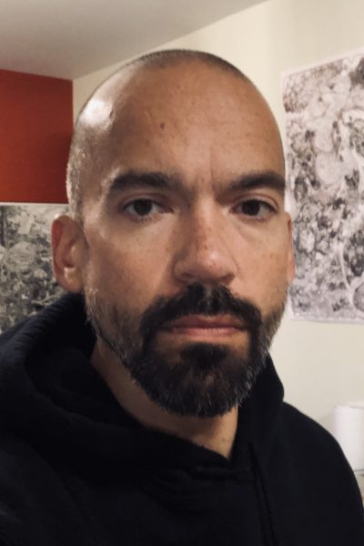 A person with a shaved head and dark beard wearing a black hoodie, photographed indoors against a white wall with red and patterned elements visible in the background.