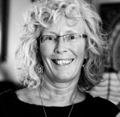 Person with curly white hair and glasses smiling at camera in black and white portrait photo.