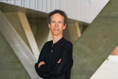 Person with curly gray hair wearing a black shirt stands with arms crossed in front of a modern concrete architectural structure.