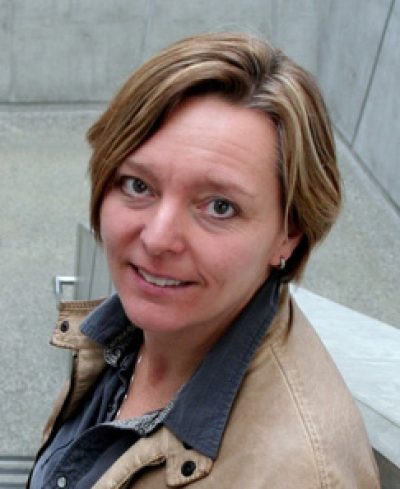 Person with shoulder-length brown hair smiling at camera, wearing tan jacket over dark shirt, against gray background.