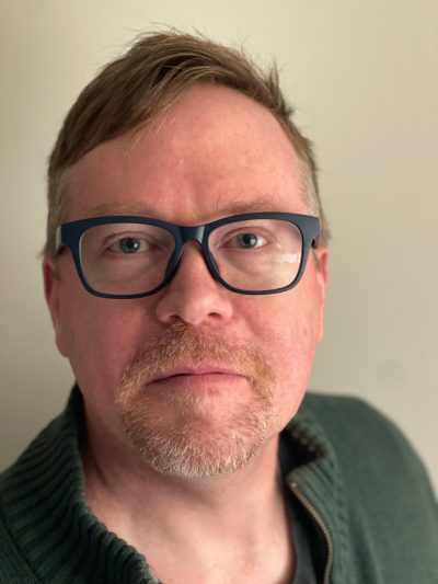 Person wearing dark-framed glasses and a green shirt, with reddish-brown hair and facial hair, looking directly at camera against a neutral background.