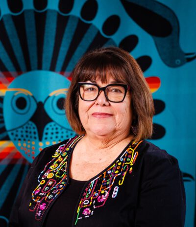 Person wearing glasses and a colorful beaded vest against a vibrant blue and black geometric patterned background.