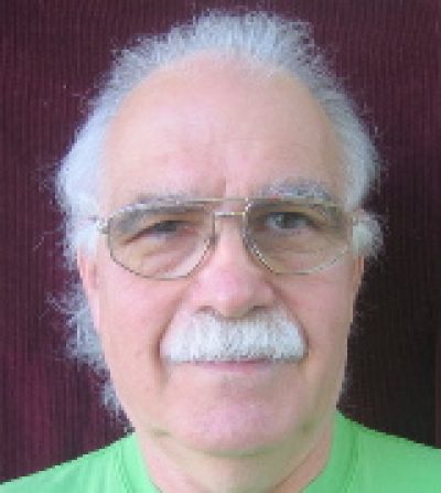 Older person with white hair and glasses wearing a green shirt, photographed against a burgundy background.