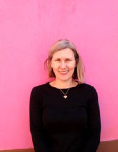 Person with blonde hair wearing a black sweater and cross necklace against a bright pink background.