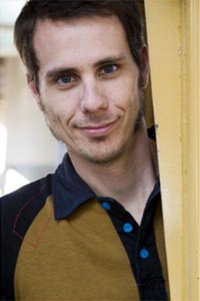 Person with dark hair wearing a mustard yellow and dark blue shirt, smiling at the camera indoors.