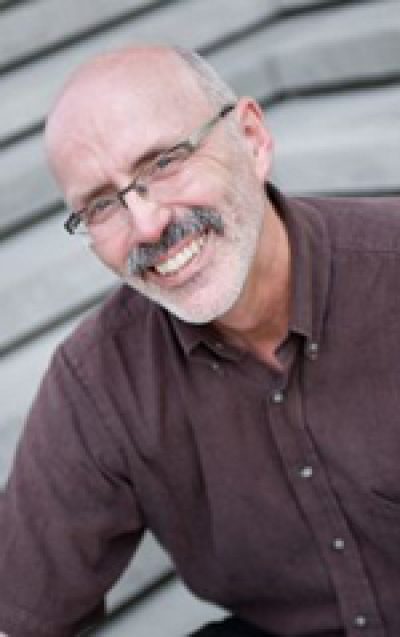 Person wearing glasses and brown shirt smiling at camera against corrugated metal background.