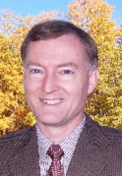 Person smiling at camera wearing brown blazer and patterned tie, with autumn foliage in background.