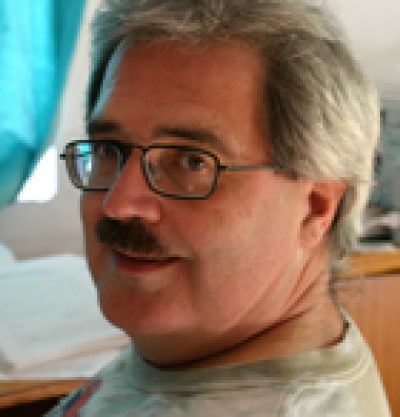 Person with gray hair and glasses wearing a gray shirt against a blue background.