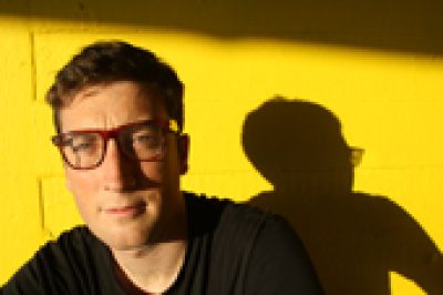 Person wearing glasses and dark clothing against a yellow background with their shadow visible beside them.
