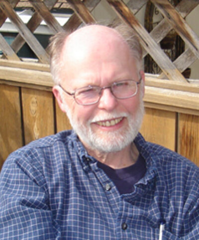 Person wearing glasses and blue plaid shirt, smiling at camera with wooden lattice and doors in background.