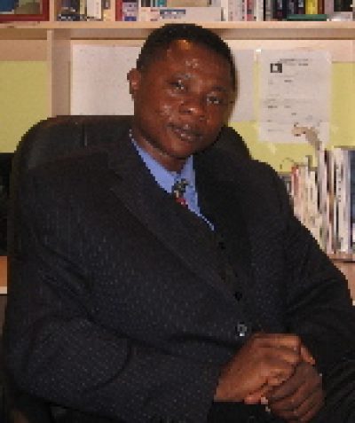 Person in dark suit and blue tie sitting indoors with bookshelves visible in background.