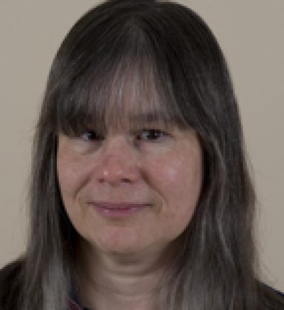 Person with dark hair and bangs, smiling slightly at the camera against a neutral beige background.