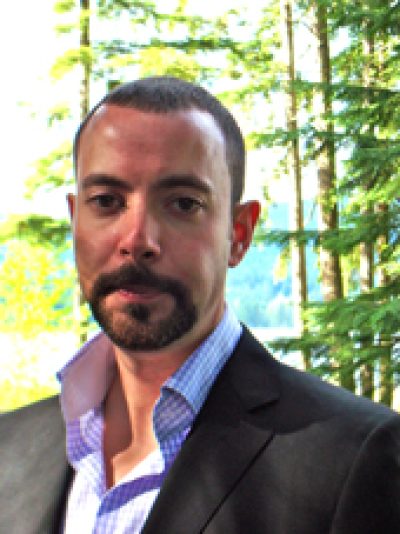 Person with dark hair and beard wearing a dark blazer and blue shirt, smiling outdoors with green foliage in background.