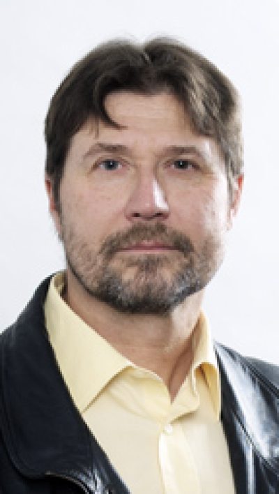 Headshot portrait of an adult person with dark hair and facial hair, wearing a cream-colored shirt and dark jacket against a light background.