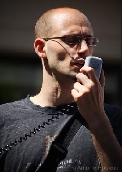 Person wearing glasses and dark clothing speaks into a microphone against a dark background.