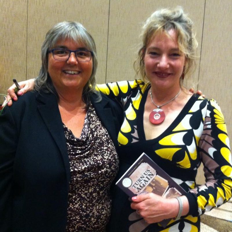 Two people posing together indoors, one wearing a black blazer and the other in a yellow and black patterned dress, holding a book.