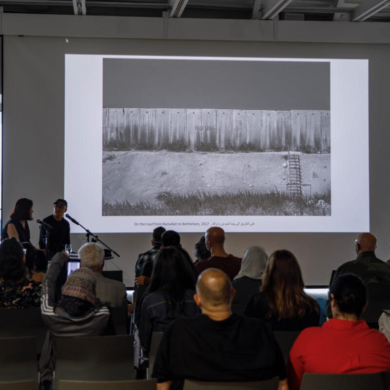 A presenter speaks to an audience in front of a large projection screen displaying a landscape photograph with text in multiple languages.