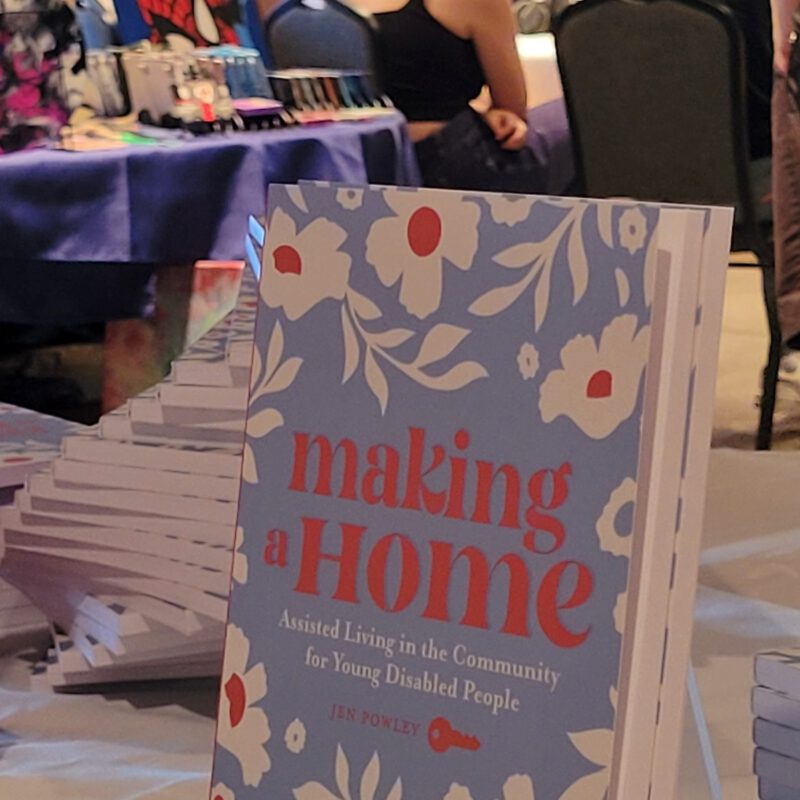 A book titled "Making a Home" displayed on a table at an indoor community event with people in the background.