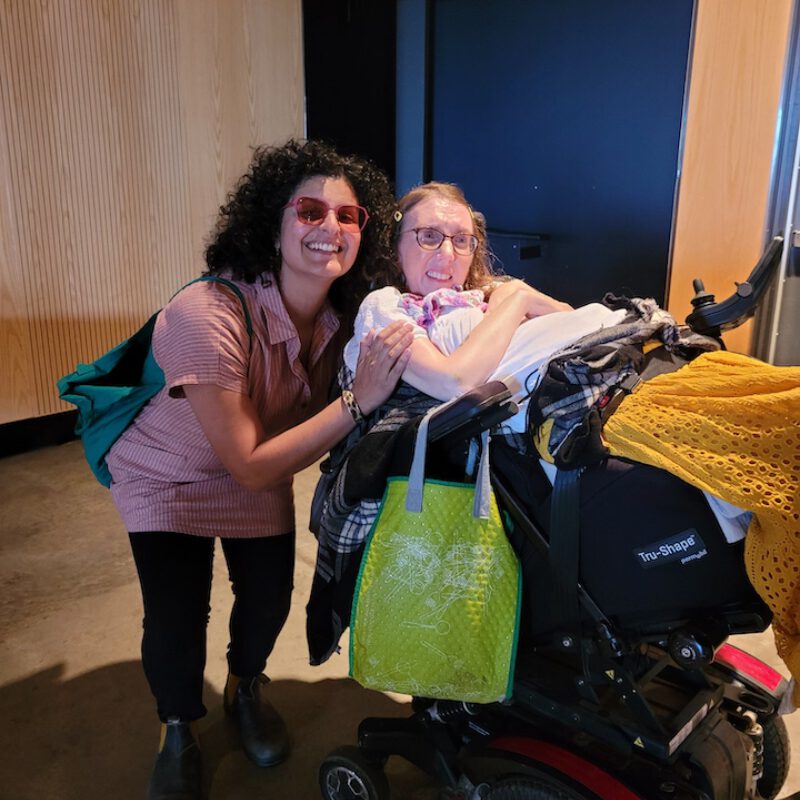 Two people smiling at a mobility scooter loaded with clothing and items in an indoor setting.